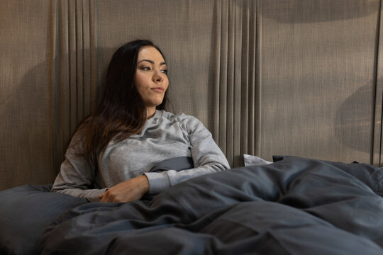 Woman reclining against pleated headboard, in gray sweatshirt, hands resting on gray duvet on bed