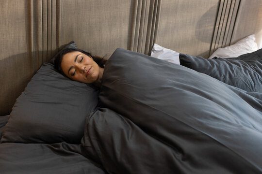 Adult Asian woman sleeping on side in bedroom under dark gray duvet with pillows