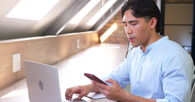 Asian man reading phone, typing on laptop, calling while in blue shirt under skylights, copy space