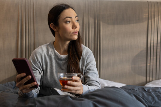 Asian woman sitting on bed holding smartphone and glass cup of tea, looking right