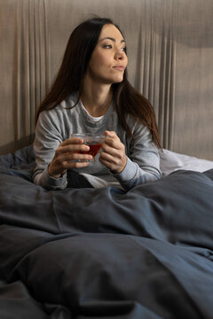 Woman sitting in bed holding glass cup of tea, wearing gray sweatshirt against pleated headboard