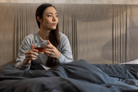 Asian woman sitting in bed wearing gray sweatshirt holding glass teacup against tufted headboard