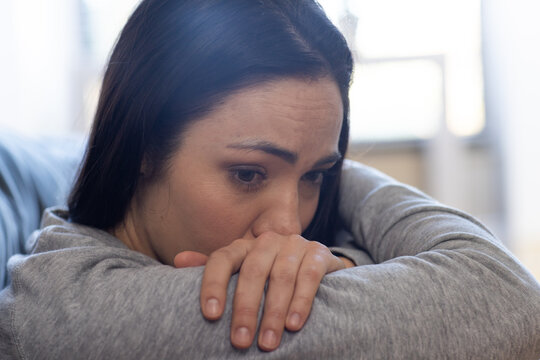 Woman wearing gray sweatshirt leaning on couch, resting chin on folded forearms under window light