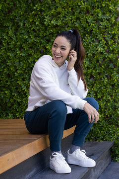 Woman sitting on bench at patio wearing hoodie, green hedge, leaning, smiling, touching scrunchie