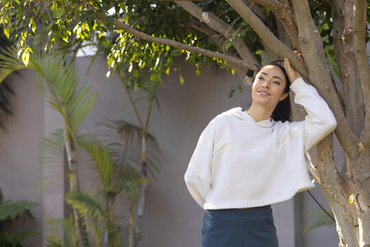 Asian woman leaning against tree in courtyard near palm plants, wearing cropped hoodie, copy space