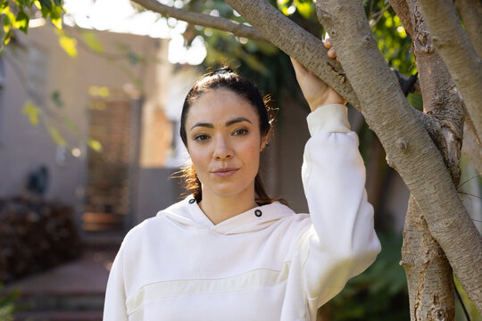 Woman standing holding tree branch and wearing white hoodie while looking at camera in backyard