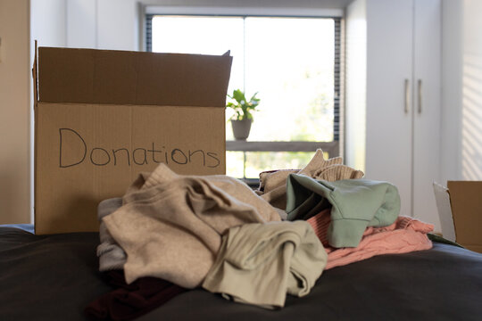 Pile of assorted clothing sitting on dark bedspread near window with donations box and plant