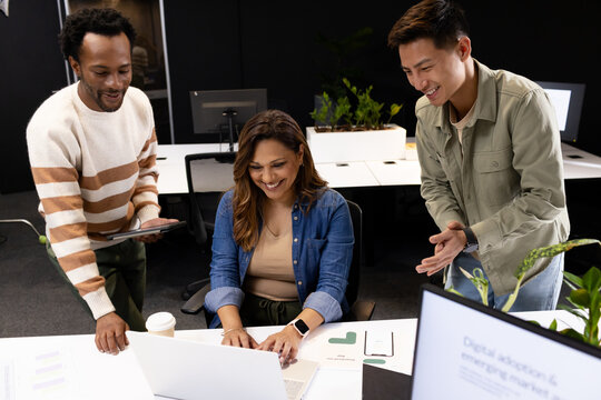 Diverse team leaning over laptop with charts in office, woman typing while peers pointing at tablet