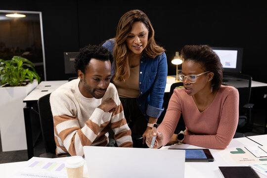 Coworkers including man in cream-and-brown striped sweater pointing at laptop and tablet on desk