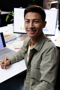 Asian man sitting at modern office workstation holding metallic pen near laptop monitor