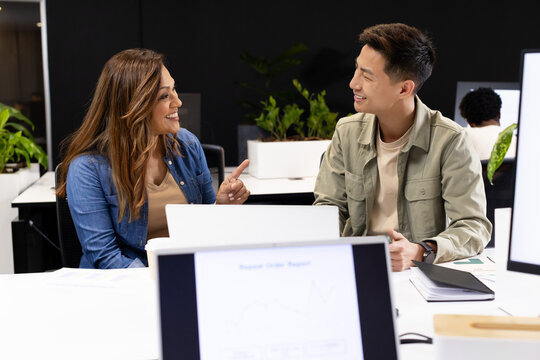 Diverse coworkers discussing project at open-plan office desk with laptops, monitor, planter