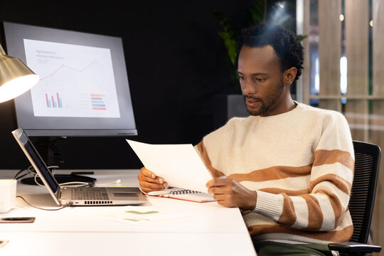 African American man sitting in office reading document while checking laptop screen, copy space