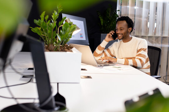 African American man in striped sweater speaking on phone, typing laptop at open office, copy space
