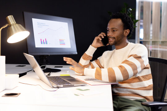 African American man talking on phone, using laptop at desk with charts on monitor, copy space