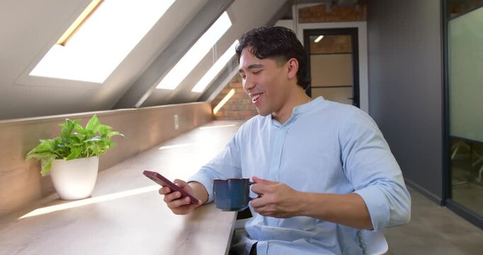 Adult man glancing at phone, scrolling, smiling, sipping mug on break at loft counter, copy space