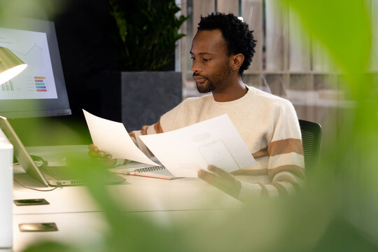 African man sitting at desk in modern office reviewing printed pages with laptop and monitor charts
