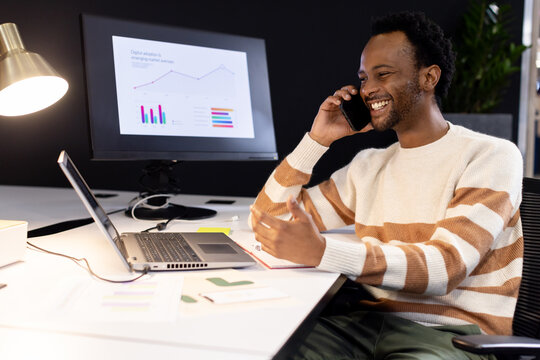 African American man talking on phone pointing at laptop and chart monitor in office, copy space