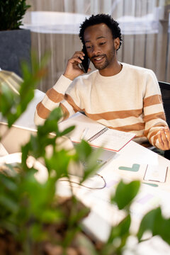 African American man in cream sweater holding phone to ear while checking laptop, notes at desk