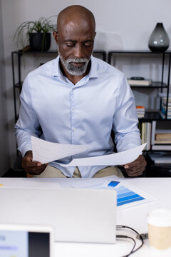 Senior man in blue shirt reading papers and color chart at office desk with laptop