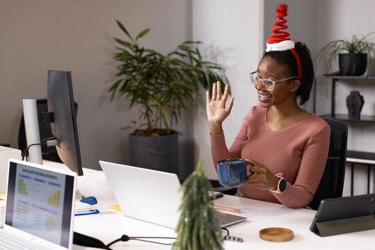 African woman wearing Santa headband holding blue mug and waving at laptop at desk, copy space