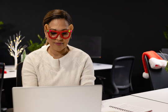 Mature African woman wearing holiday glasses, working on silver laptop at office desk with notebook