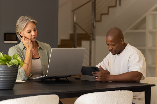 Diverse couple sitting at dining table at home using laptop and tablet with planter