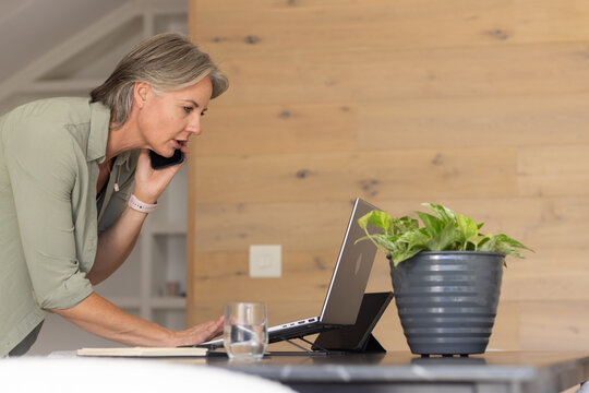 Mid-40s woman in green top at home leaning, holding phone, operating laptop on stand, copy space