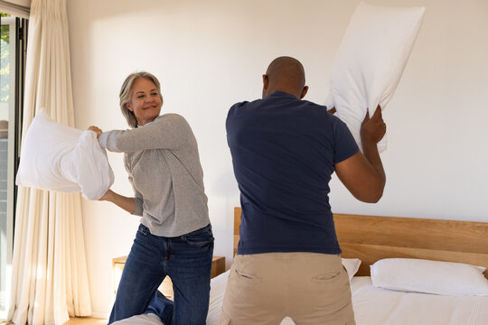 Couple playing pillow fight on made bed in bright bedroom with white pillows and curtains