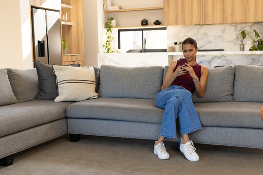 African American woman sitting on right side of gray L-shaped sofa scrolling smartphone, copy space