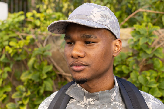 Male standing and looking to right by stone wall with vines, wearing camo uniform, backpack straps