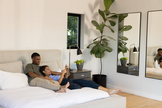 African American couple reclining in bedroom on bed with white duvet, woman holding smartphone