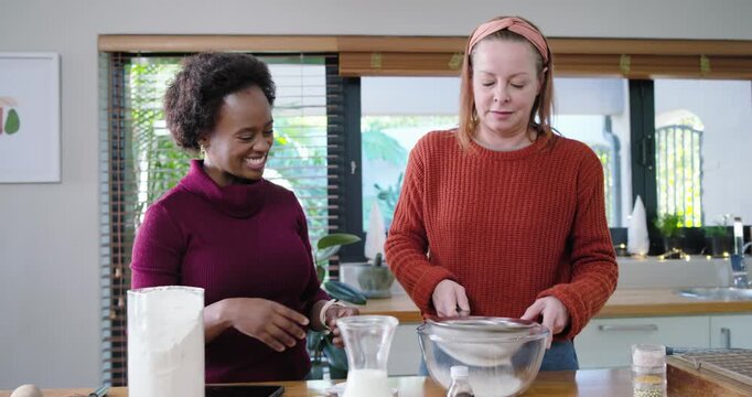 Diverse female pair measuring flour, sifting with sieve into glass bowl at counter, making batter