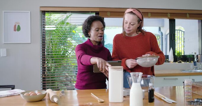Diverse female friends following tablet recipe while measuring flour into glass bowl for baking
