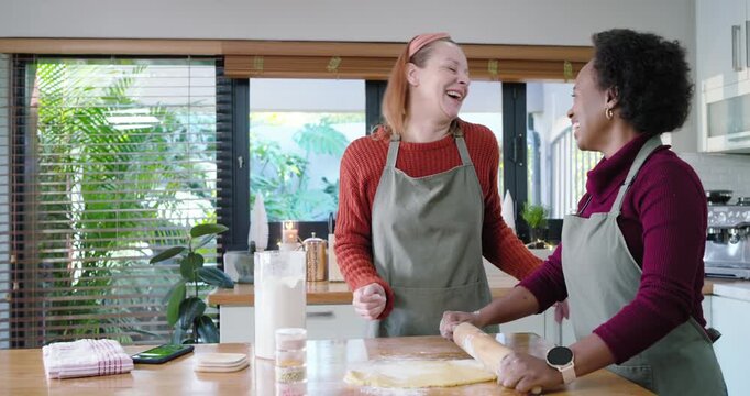 Two women wearing aprons sprinkling flour, laughing and rolling dough at kitchen with rolling pin
