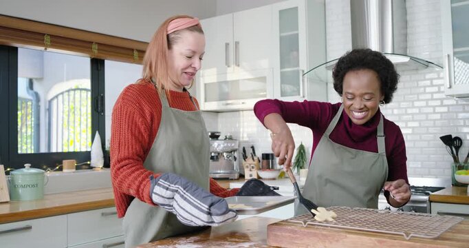 Diverse female friends holding hot tray with mitt, sliding cookies onto cooling rack with spatula