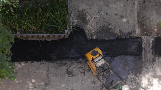 Construction worker compacting fresh asphalt with vibrating plate compactor on sidewalk. Top view of road maintenance worker repairing pavement and creating new asphalt surface outdoors