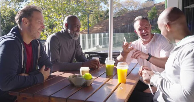 Diverse male friends laughing on sunny patio while rightmost man fiddling straw lid to entertain