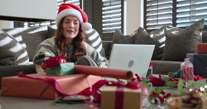 Female sitting on sofa wearing Santa hat, noticing open laptop, showing gift toward screen