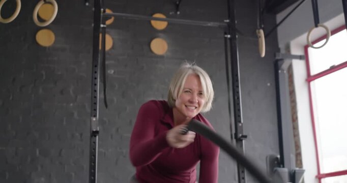 Senior woman in maroon top, leggings, gripping battle ropes, making rope waves for strength at gym