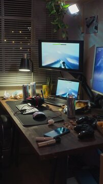 Vertical video of messy gaming desk with screens displaying active shooter game at night. Clip is ideal for projects on screen addiction, gaming lifestyle, or youth technology habits