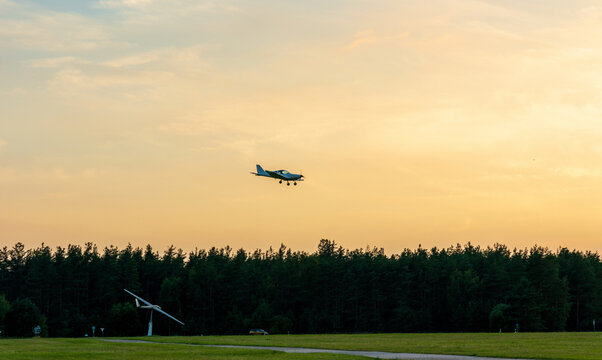 View of a light aircraft approaching the runway at Paluknys Airfield with a glider on display in the foreground and a dense forest under a golden sunset sky in Paluknys, Vilnius County, Lithuania.