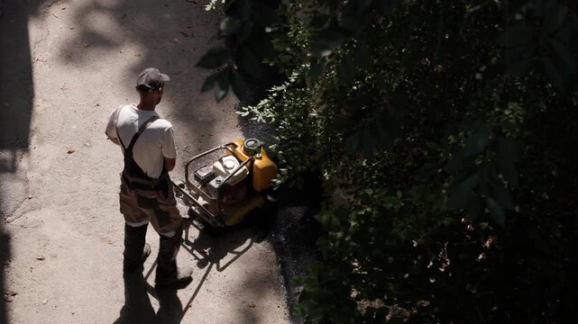 Construction worker compacting fresh asphalt with vibrating plate compactor on sidewalk. High angle view of road maintenance worker repairing and resurfacing pedestrian pavement outdoors