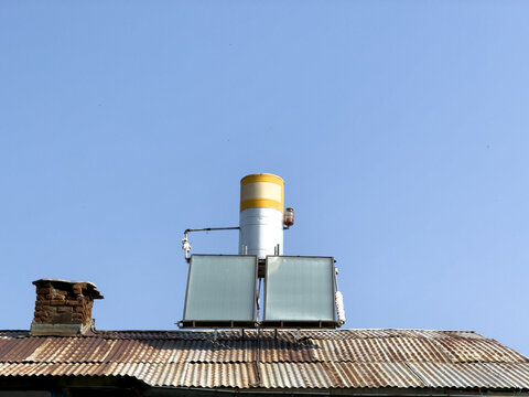 View of a solar water heater system mounted on a corrugated metal roof with a brick chimney against a clear blue sky in Posof, Ardahan, Turkiye.