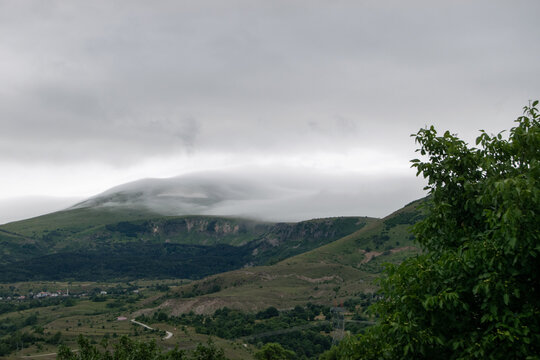 View of the mountainous landscape of Posof with low-hanging clouds over the green hills and a small settlement in the distance in Posof, Ardahan, Turkiye.