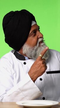 Indian Senior Male Sikh Chef in Traditional Attire Sitting at Table, Pointing at Empty Plate and Showing OK Sign on Green Screen