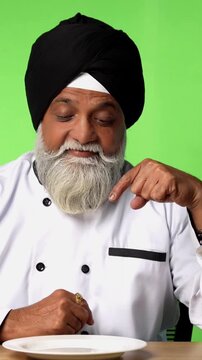 Indian Senior Male Sikh Chef in Traditional Attire Sitting at Table, Pointing at Empty Plate and Showing OK Sign on Green Screen