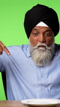 Indian Asian Senior Male Sikh Chef Sitting Isolated at Table with Empty Plate and Hand Fan Made of Indian Rupee Notes on Green Screen Background