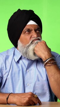 Indian Asian Senior Sikh Man in Casual Clothing Sitting Isolated at Table on Green Screen, Showing Surprise Expressions, Thinking, Thumbs Up, or Pointing at Blank Space
