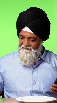 Indian Asian Senior Sikh Man in Casual Clothing Sitting Isolated at Table on Green Screen, Holding Spoon and Fork with Excitement Above Empty Dinner Plate