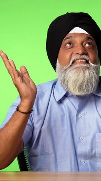 Indian Asian Senior Sikh Man in Casual Clothing Sitting Isolated at Table on Green Screen, Showing Surprise Expressions, Thinking, Thumbs Up, or Pointing at Blank Space